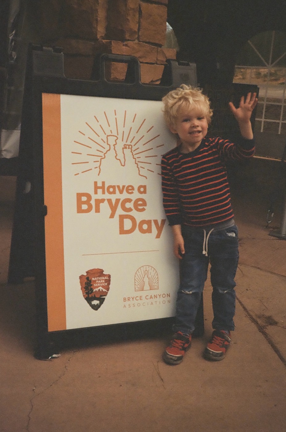 Bryce waving next to a sandwich-board sign at Bryce Canyon that reads Have a Bryce Day